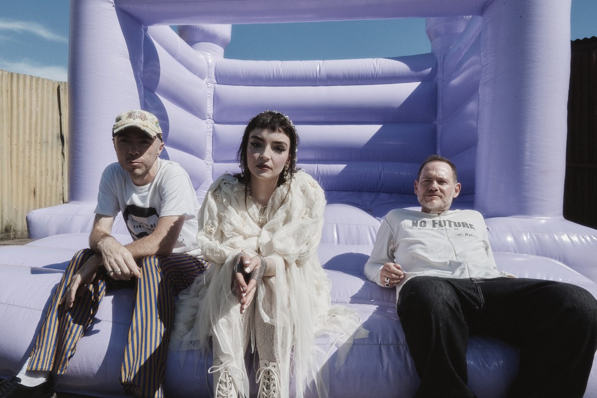 Die drei Mitglieder von CHVRCHES sitzen vor einer hellvioletten Hüpfburg